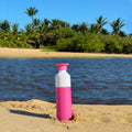 Dopper Insulated Small Pelican Pink( Thermosfles roze) - rechtop staand in zand aan strand met palmbomen op achtergrond