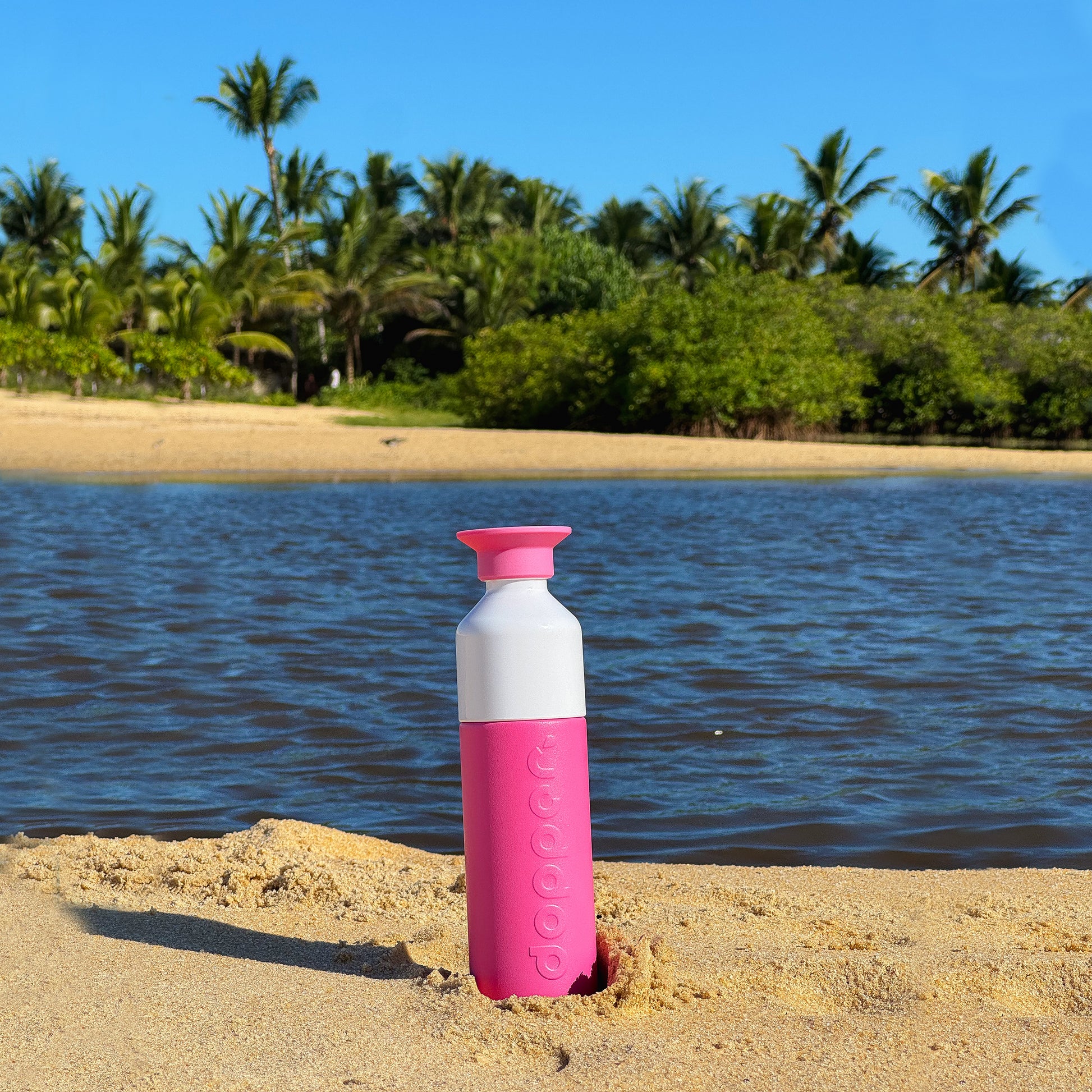 Dopper Insulated Small Pelican Pink( Thermosfles roze) - rechtop staand in zand aan strand met palmbomen op achtergrond