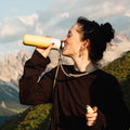 Insulated Bottle 350 ml Maple Yellow (yellow) - woman drinking from the bottle with mountains behind