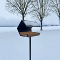 Vogelvoederhuis Open Diamond met Paal in donkergrijs in sneeuwlandschap met bomen op achtergrond