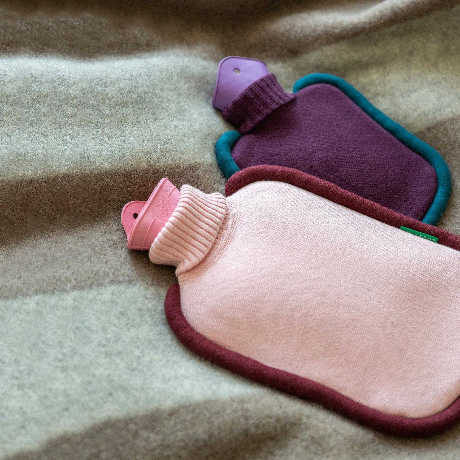 Warmwaterkruik Bumper roze en mini versie paars naast elkaar liggend op grijze deken