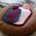 Warwaterkruik Bumper Pink en Mini versie Aubergine (paars) van Suite702 liggend op rond bruin kussen