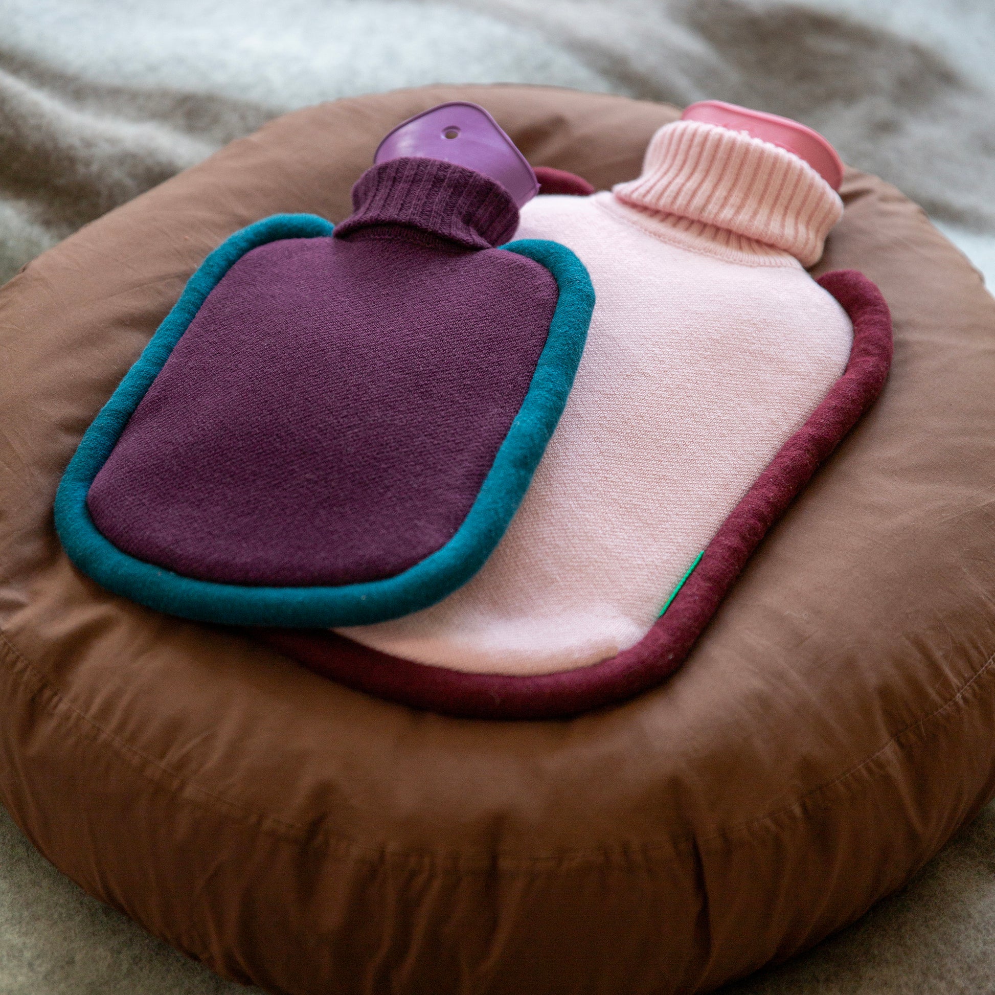 Warwaterkruik Bumper Pink en Mini versie Aubergine (paars) van Suite702 liggend op rond bruin kussen