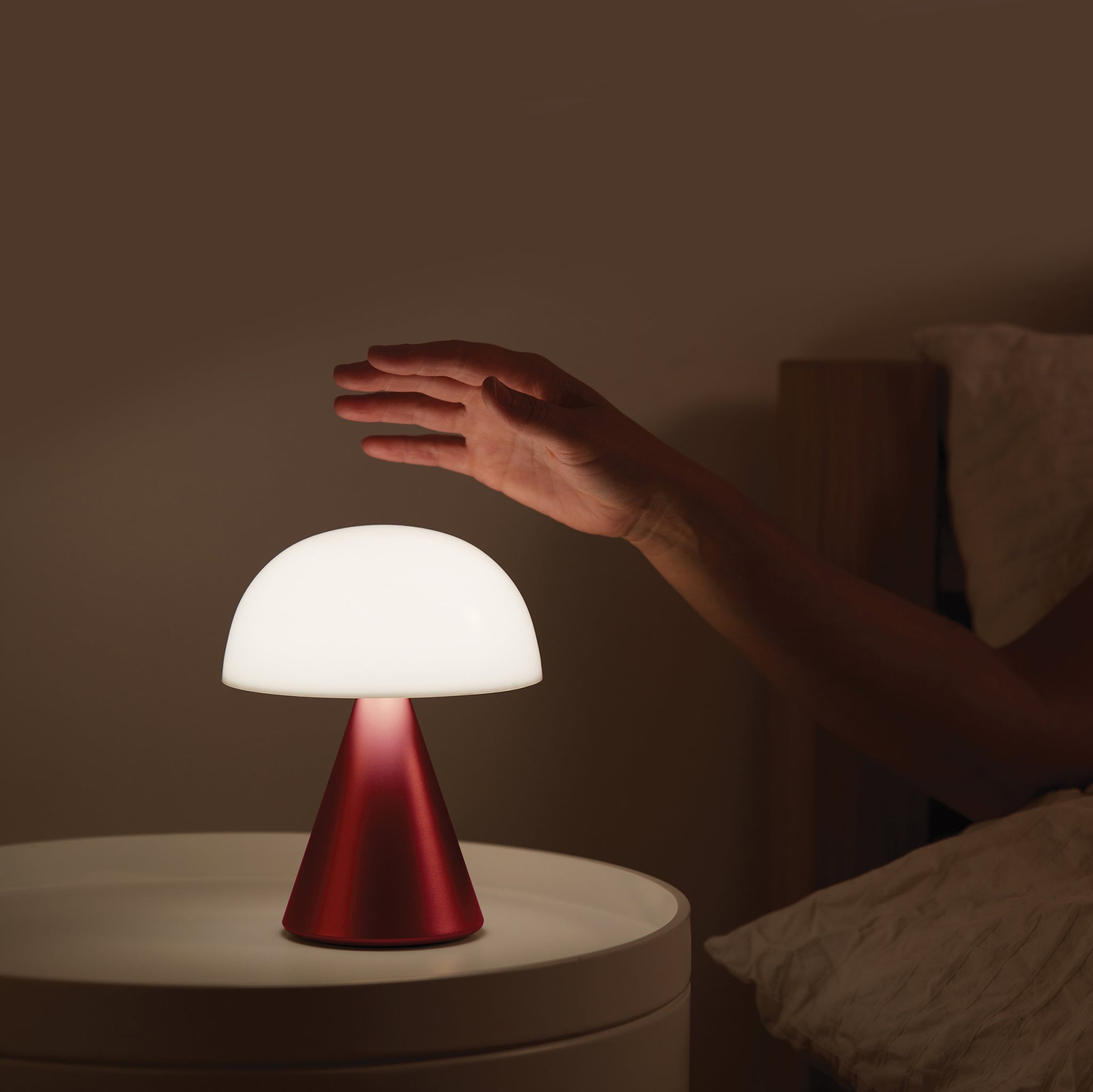 Lexon Mina Large Oplaadbare Tafellamp Donkerrood│art. LH65DR│naast bed op nachtkastje met hand boven licht