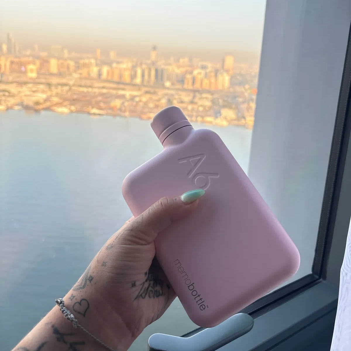 Vrouwenhand met tatoeage houdt Memobottle Waterfles A6 Steel roze voor raam met skyline op achtergrond.