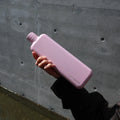 Memobottle Slim flat stainless steel water bottle in pink in a woman's hand in front of a concrete wall.