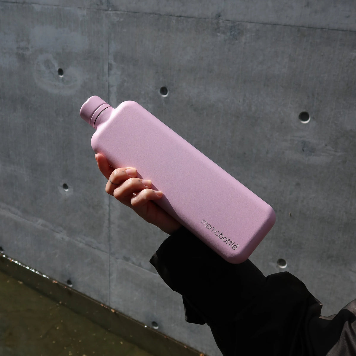 Memobottle Slim flat stainless steel water bottle in pink in a woman's hand in front of a concrete wall.