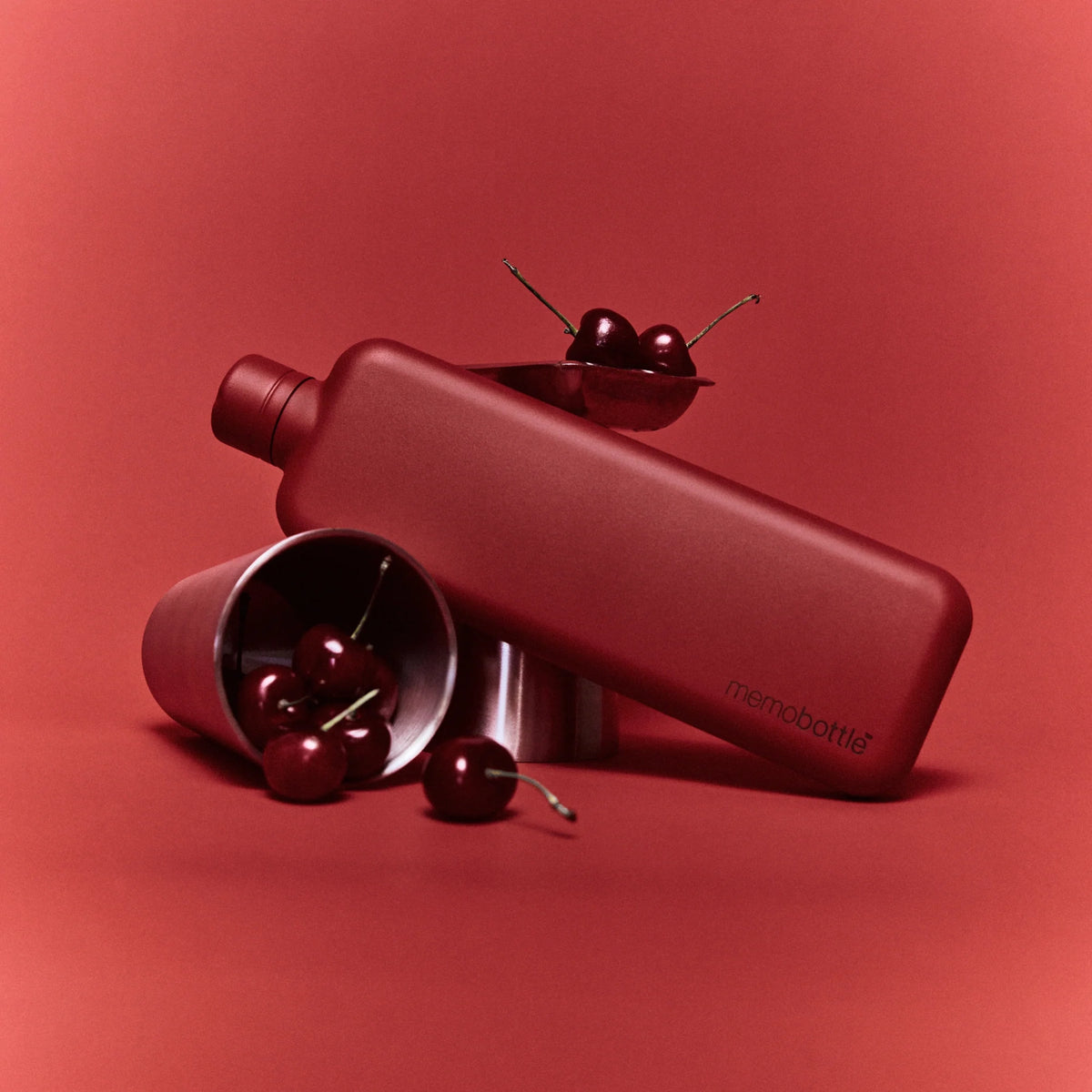 Memobottle Slim platte waterfles RVS rood, liggend op een beker met kersen voor een rode achtergrond.