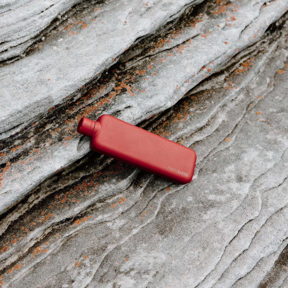 Memobottle Slim platte waterfles RVS rood, liggend op een stenen ondergrond in de natuur.