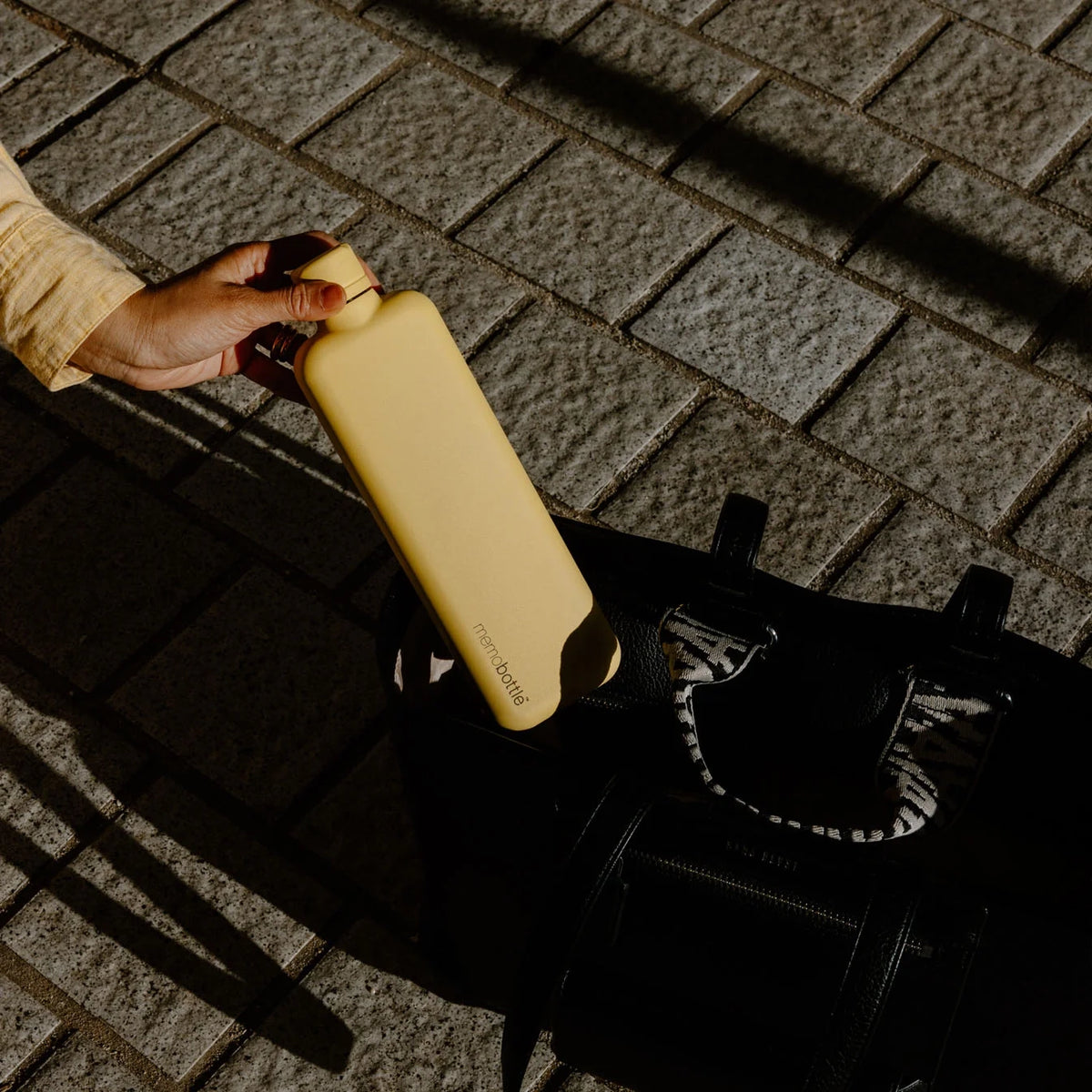 Woman putting the Memobottle Slim flat stainless steel water bottle in yellow into her bag outdoors.