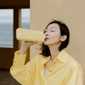 Woman in a yellow top drinking from the Memobottle Slim flat stainless steel water bottle in yellow.