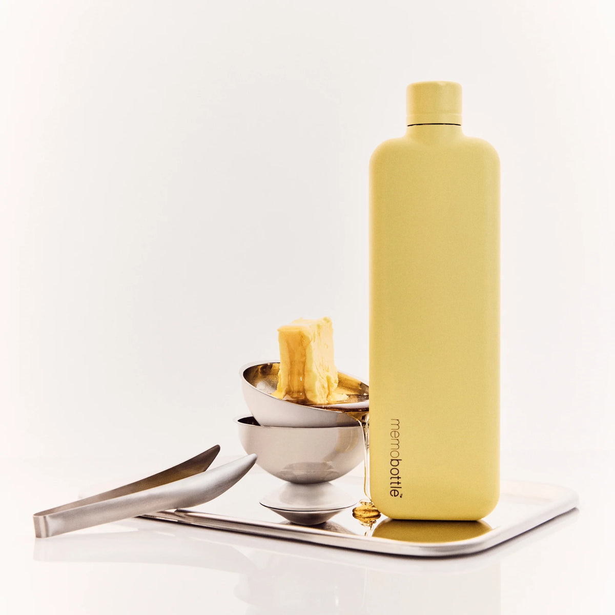 Memobottle Slim flat stainless steel water bottle in yellow on an iron tray with small bowls and a pair of tongs.