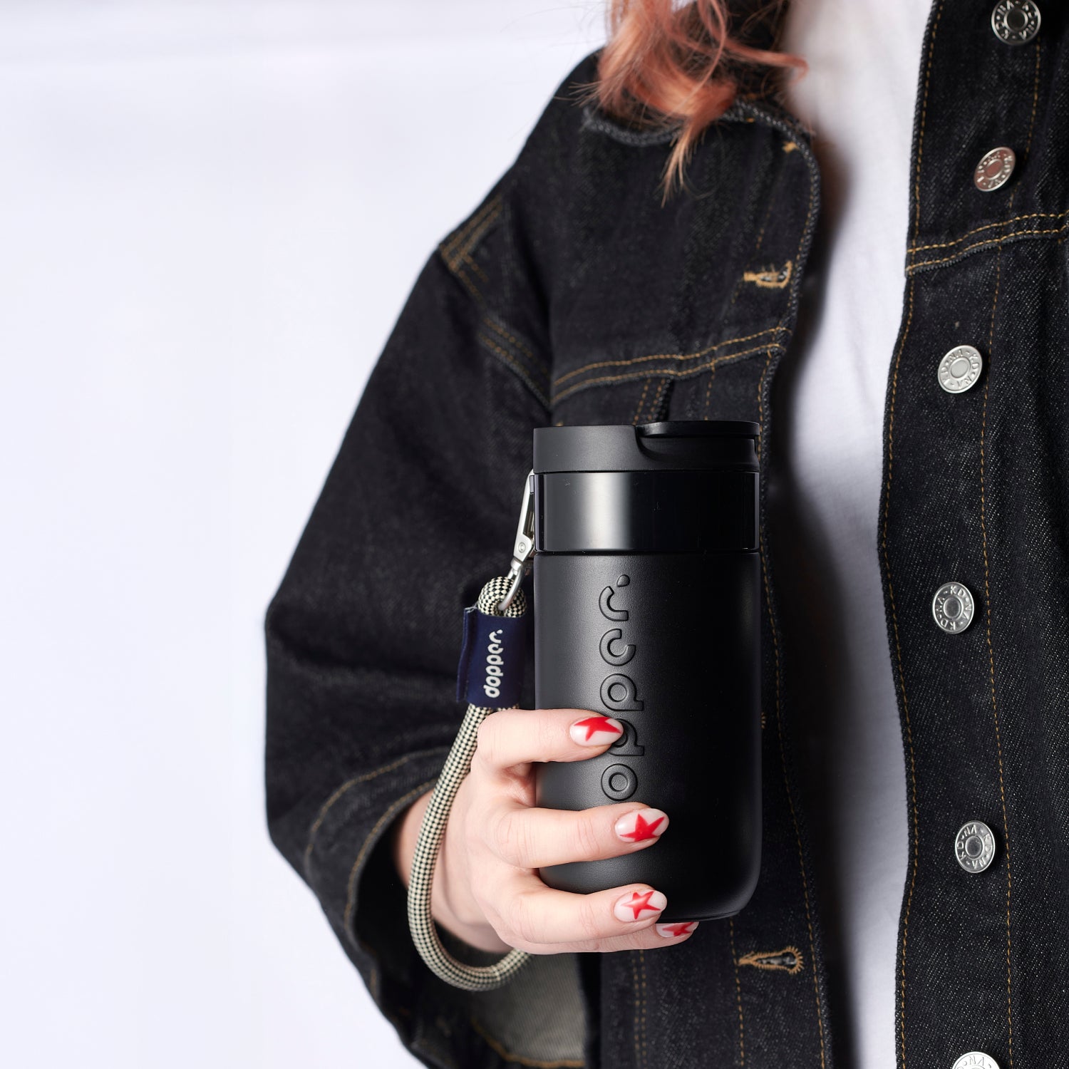 Vrouw met spijkerjack houdt Dopper Travel Mug Blazing Black met Wrist Cord Sandy lack om pols in hand