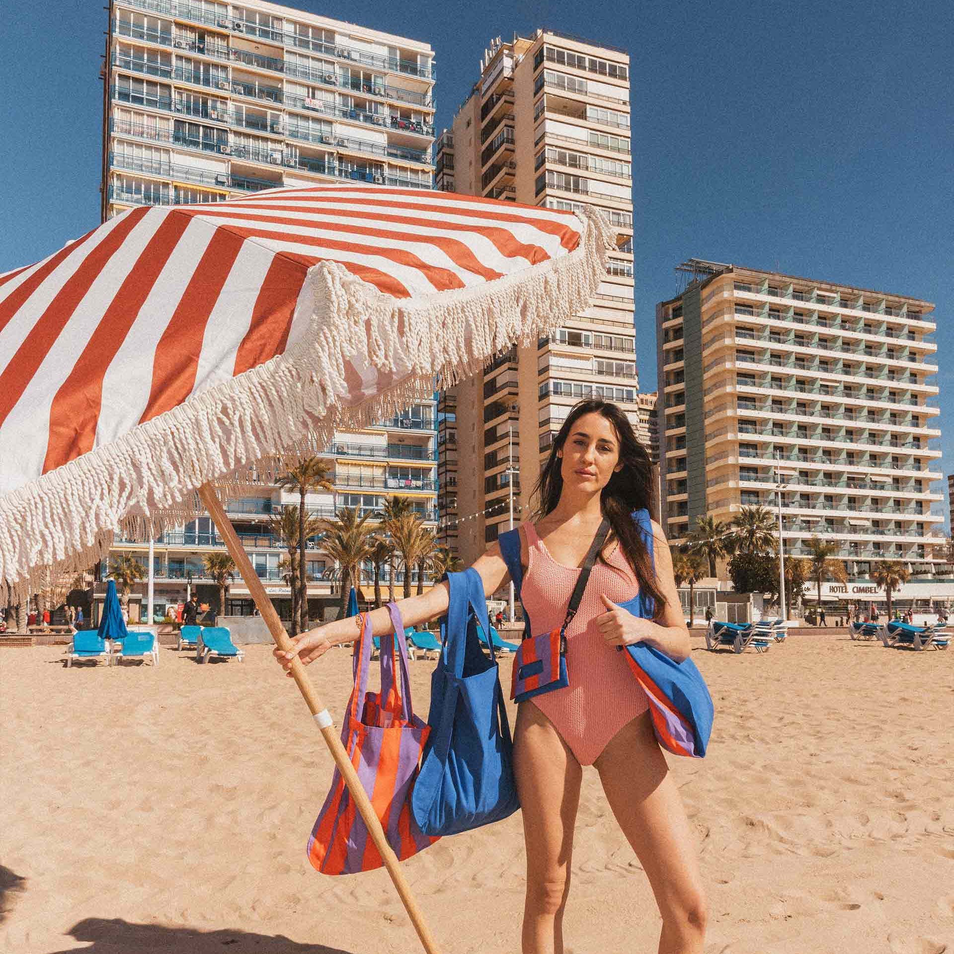 Vrouw op het strand met parasol en Notabag Red & Purple Stripes over de arm