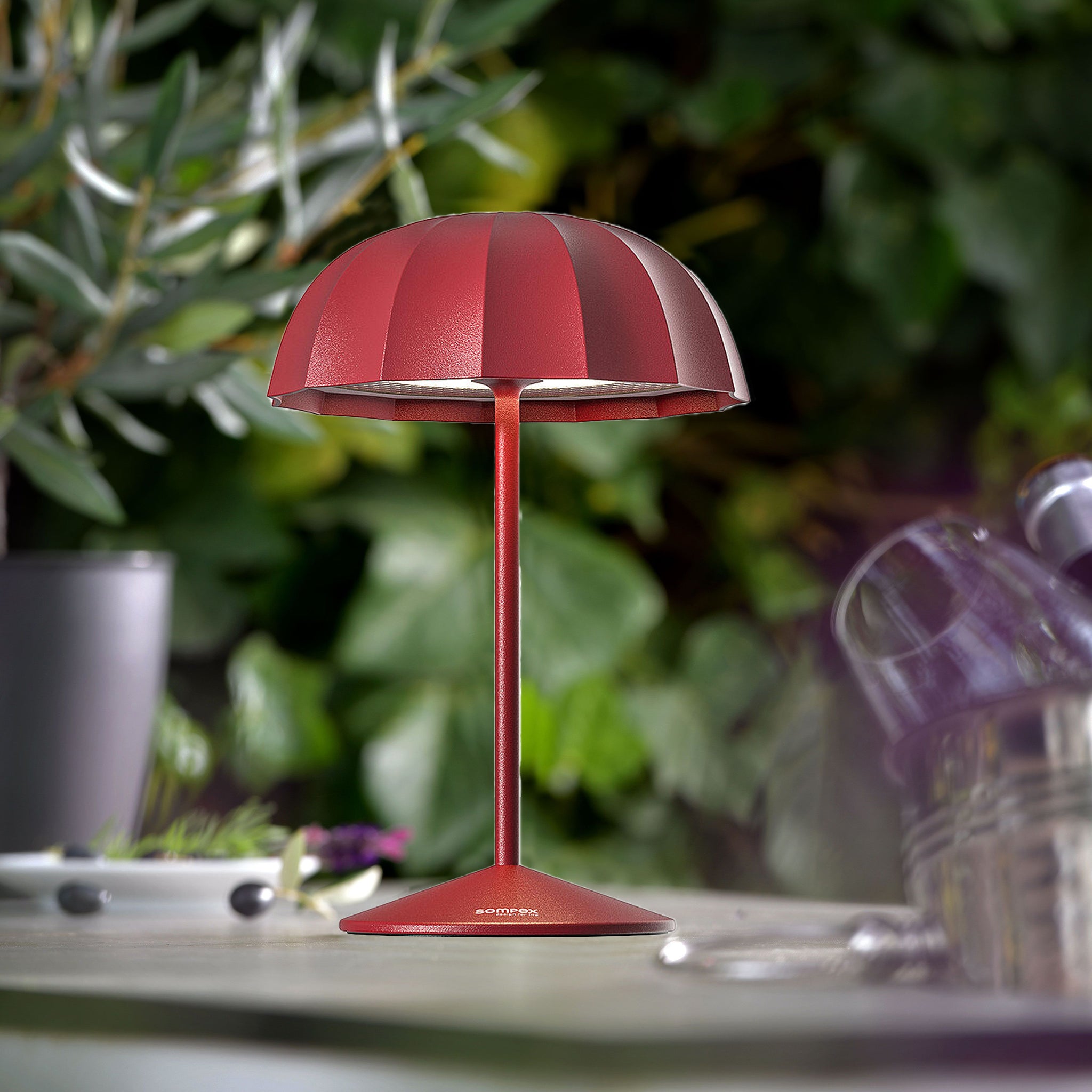 Sompex Ombrellino Oplaadbarre Buitenlamp│Rood│art. 78354│op buitentafel met struiken op achtergrond