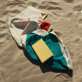 Strandlaken Martens & Martens Suite702│art. CMMTT90PITULIRE│bovenaanzicht opgevouwen op strand met boekje
