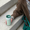 Vrouw op betonnen trap houdt Dopper Travel Mug Small 300ml Forest Green in hand