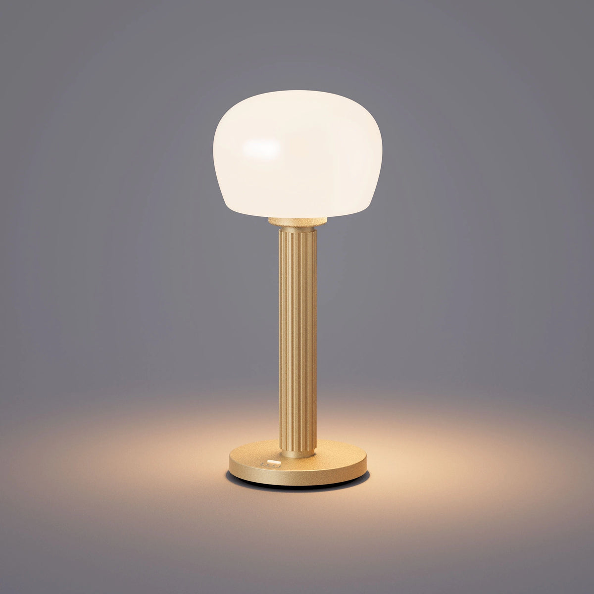 ELLE Decoration Vendome table lamp in matt gold with the light on, against a grey background.