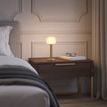 ELLE Decoration Vendome table lamp in matt gold on a bedside table next to a bed.