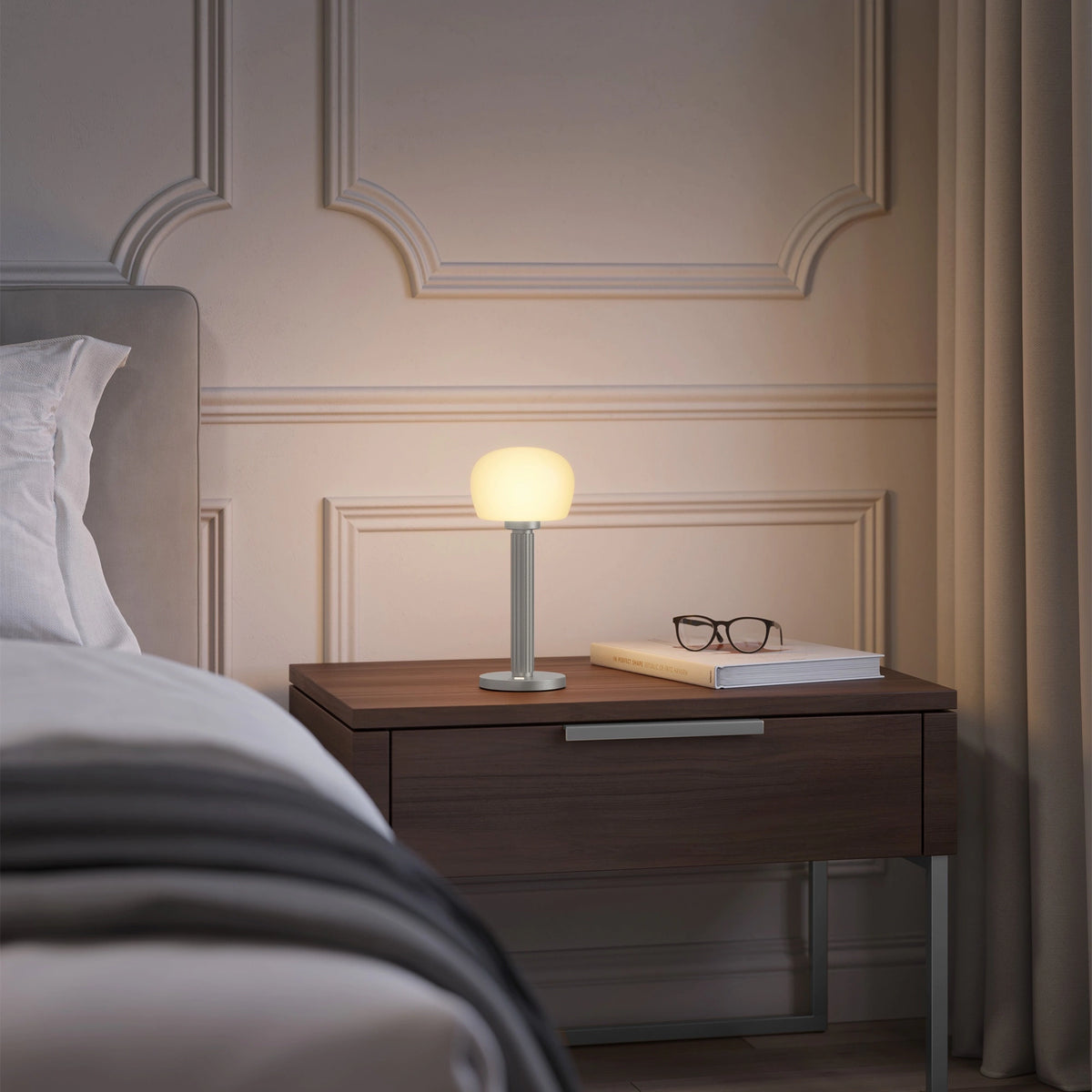 ELLE Decoration Vendome Tischleuchte in Mattsilber auf einem Nachttisch neben einem Bett.