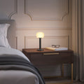ELLE Decoration Vendome table lamp in matt black with the light on, on a bedside table next to a bed.
