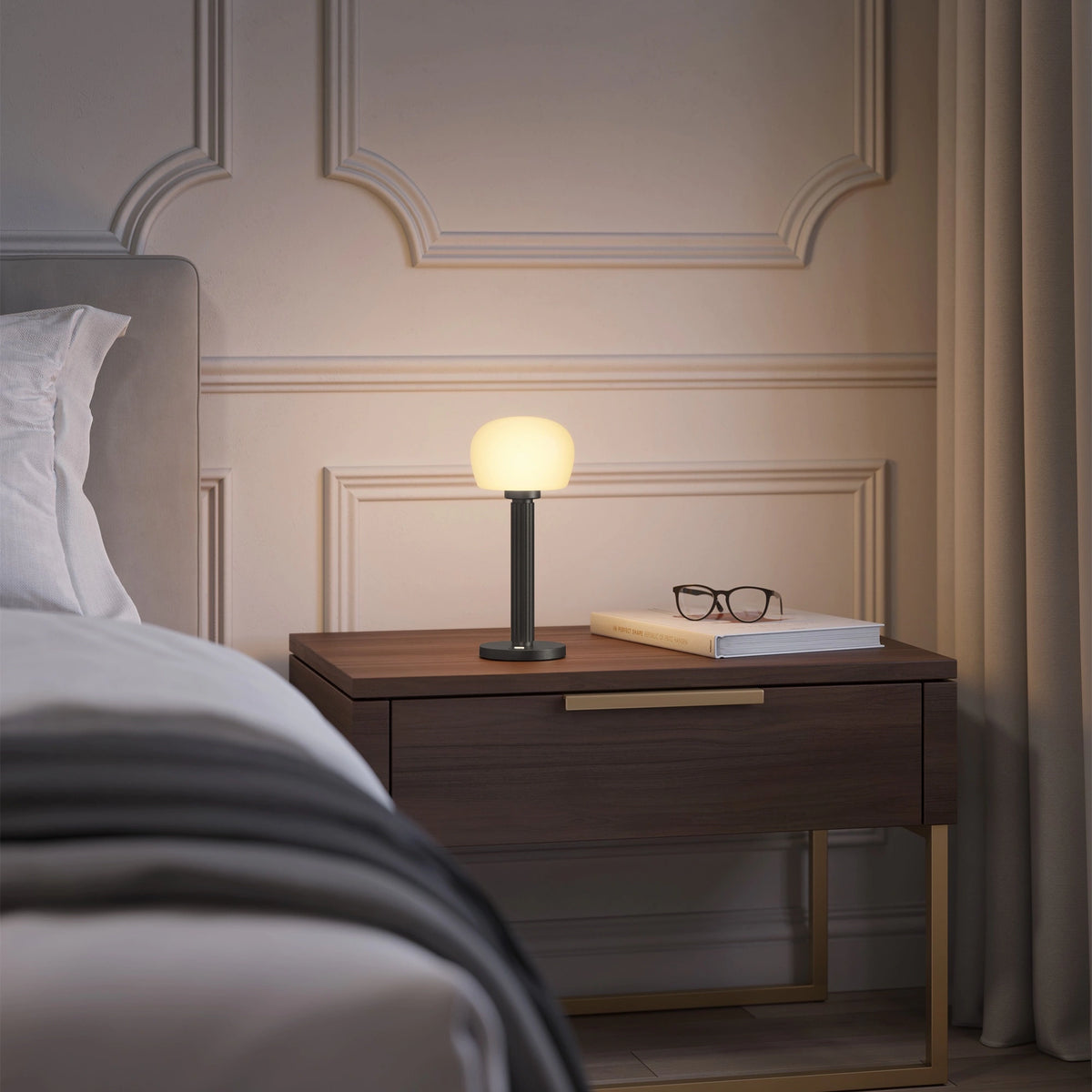 ELLE Decoration Vendome table lamp in matt black with the light on, on a bedside table next to a bed.